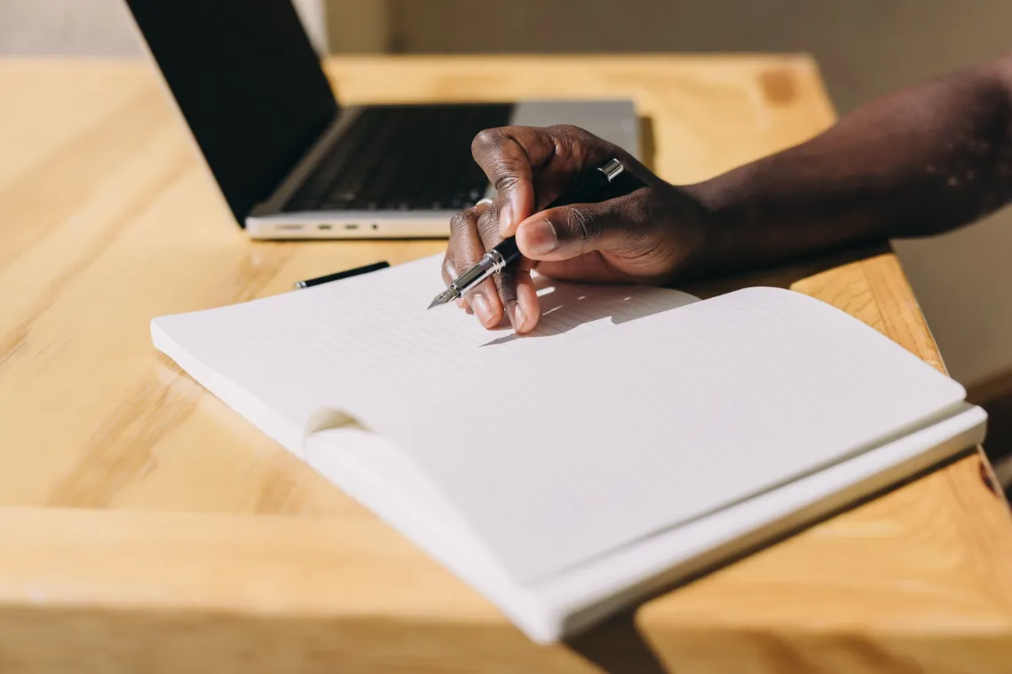 Woman's hands writing in an open notebook with an open laptop next to it.