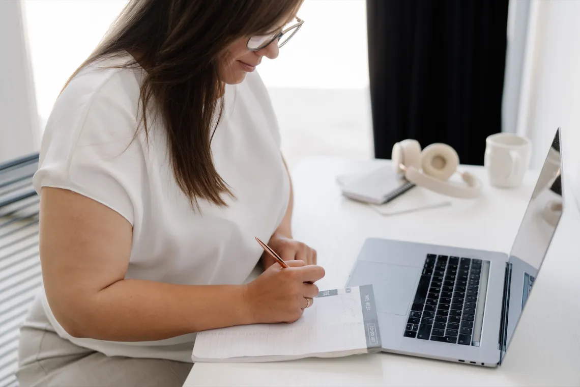 A businesswoman sitting at the desk with a laptop and a notepad, outlining a website planning process.