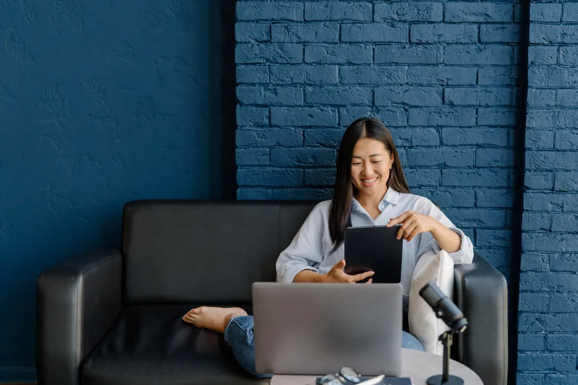 Businesswoman sitting on couch looking at a tablet with laptop and microphone on a coffee table.