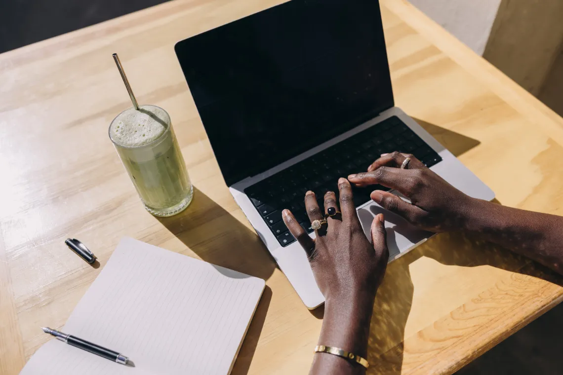 Woman's hands typing on a laptop, a notebook, pen and matcha latte nearby.