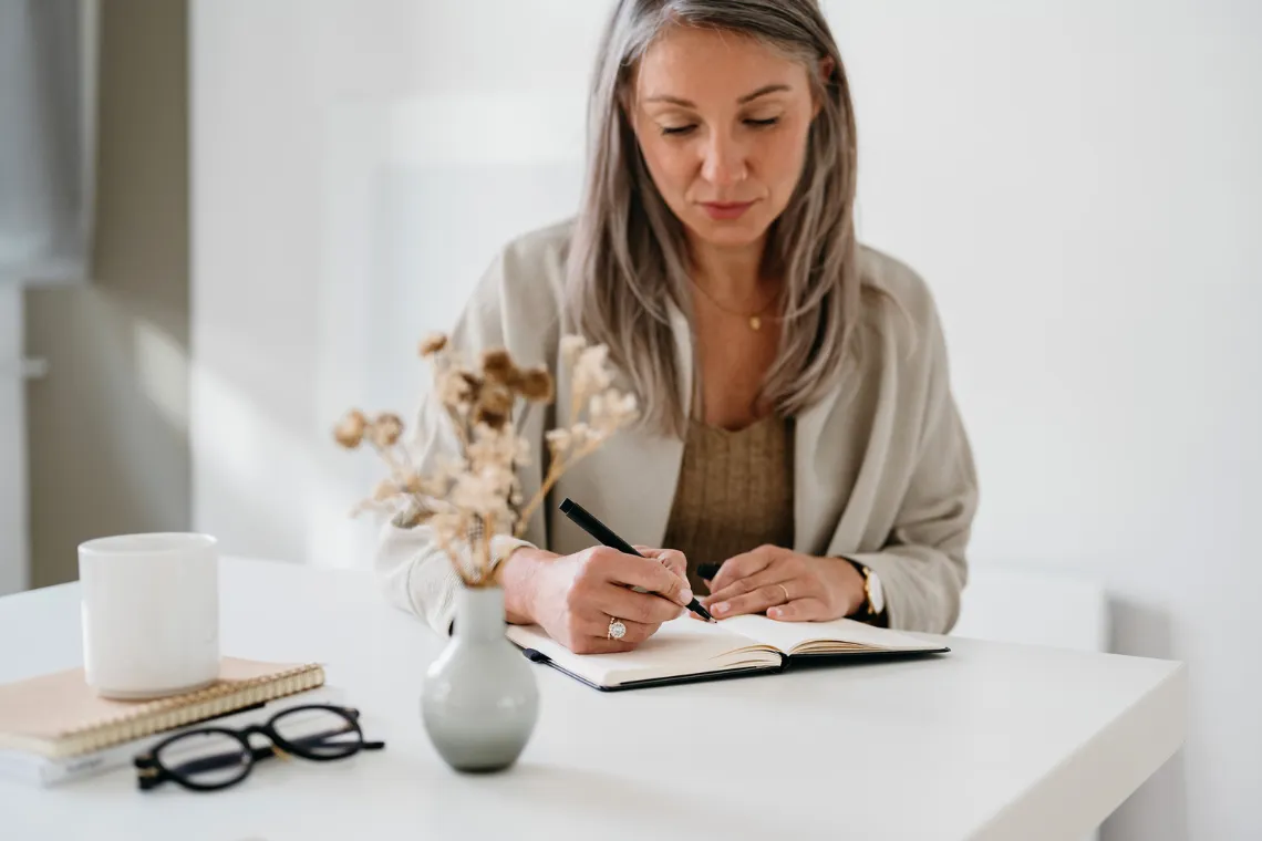 Woman sitting at a desk, writing in a notebook, flowers and coffee nearby.