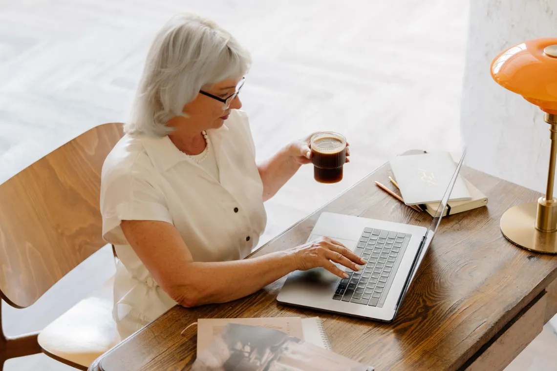 A woman sitting at a table with an open laptop in front of her, holding a cup of coffee.