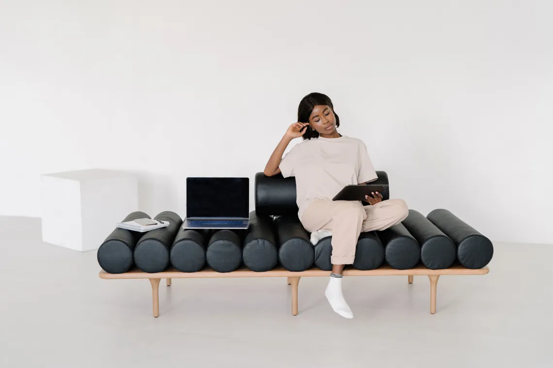 A woman sitting on a couch with a tablet in hand, open laptop sitting on the couch next to her.