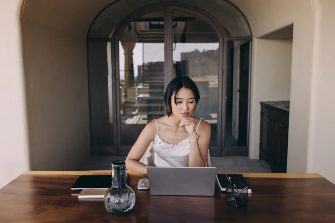 Woman sitting at a table behind an open laptop, with notebooks nearby.