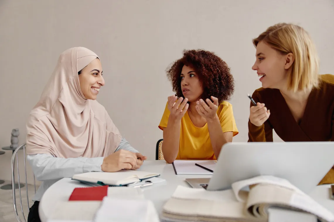 Three businesswomen sitting at a table talking with notebooks and a laptop nearby