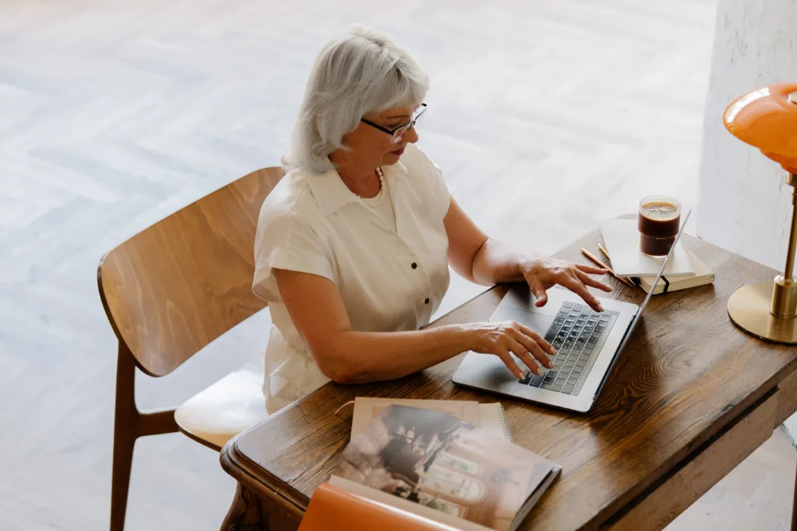 A woman sitting at a desk typing on a laptop, coffee and notebooks nearby.