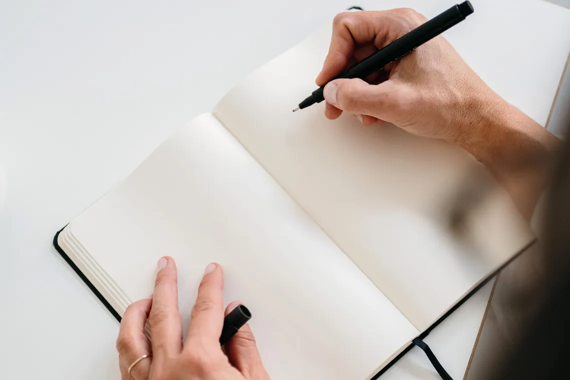 Empty notebook open on desk and woman's hands holding a pen, prepared to plan a sustainable marketing strategy.
