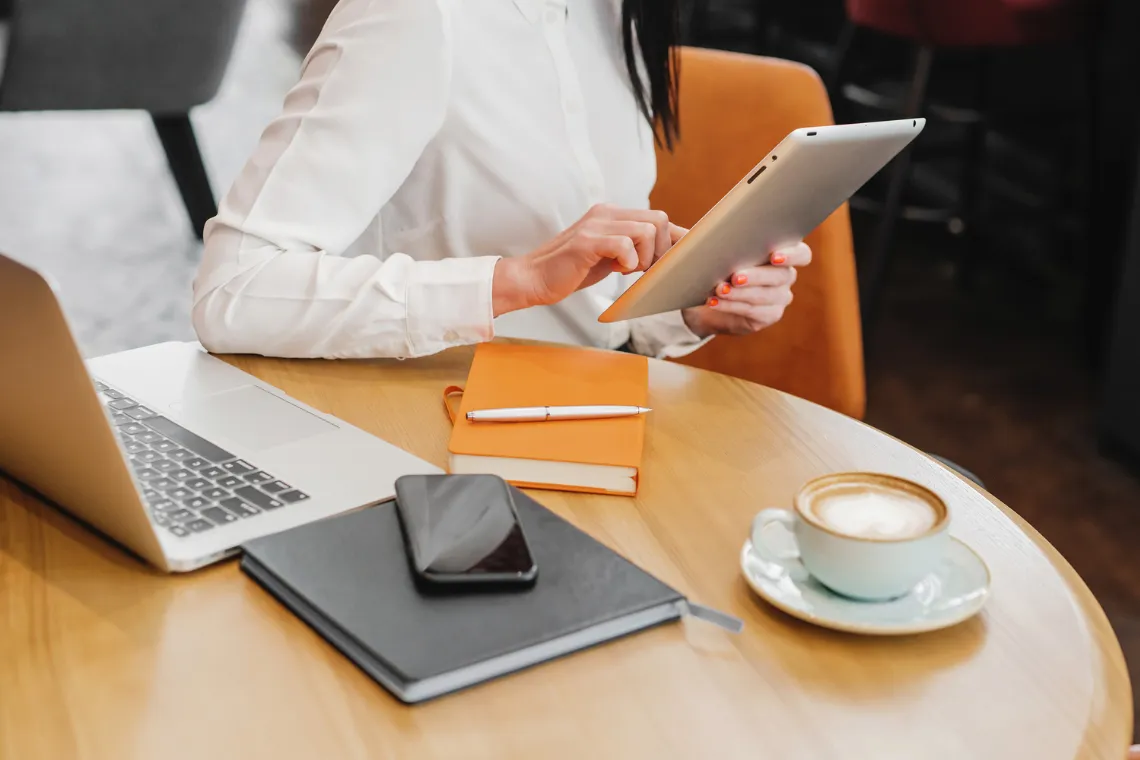 A woman sitting at a table with tablet in hand, laptop, notebook, pen and phone sit on the table next to a cup of coffee.