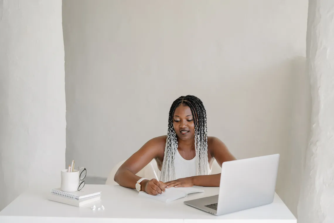 Woman sitting at a desk writing in a notebook, open laptop and notebooks on desk nearby.