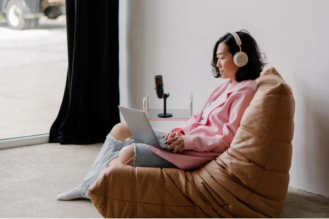 A woman sitting in a lounge chair with a laptop on her lap, headphones on, microphone on the side table.