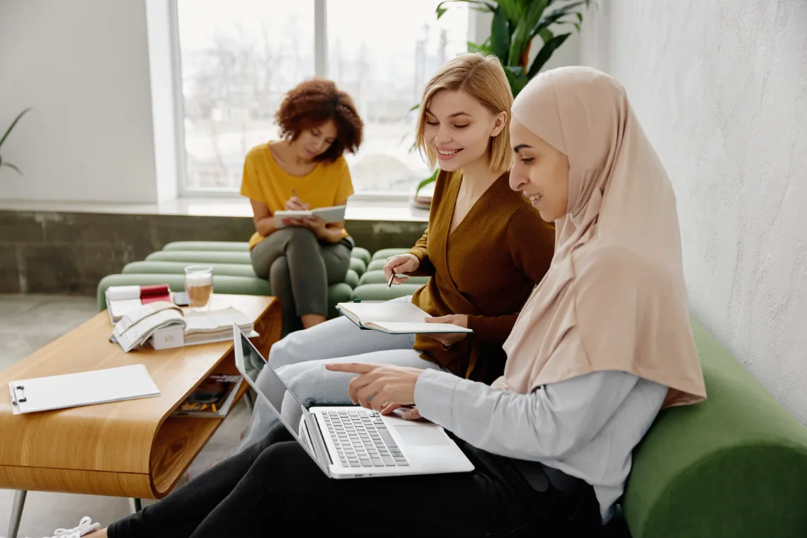 Three businesswomen sitting on a couch, writing in notebooks and typing on a laptop.