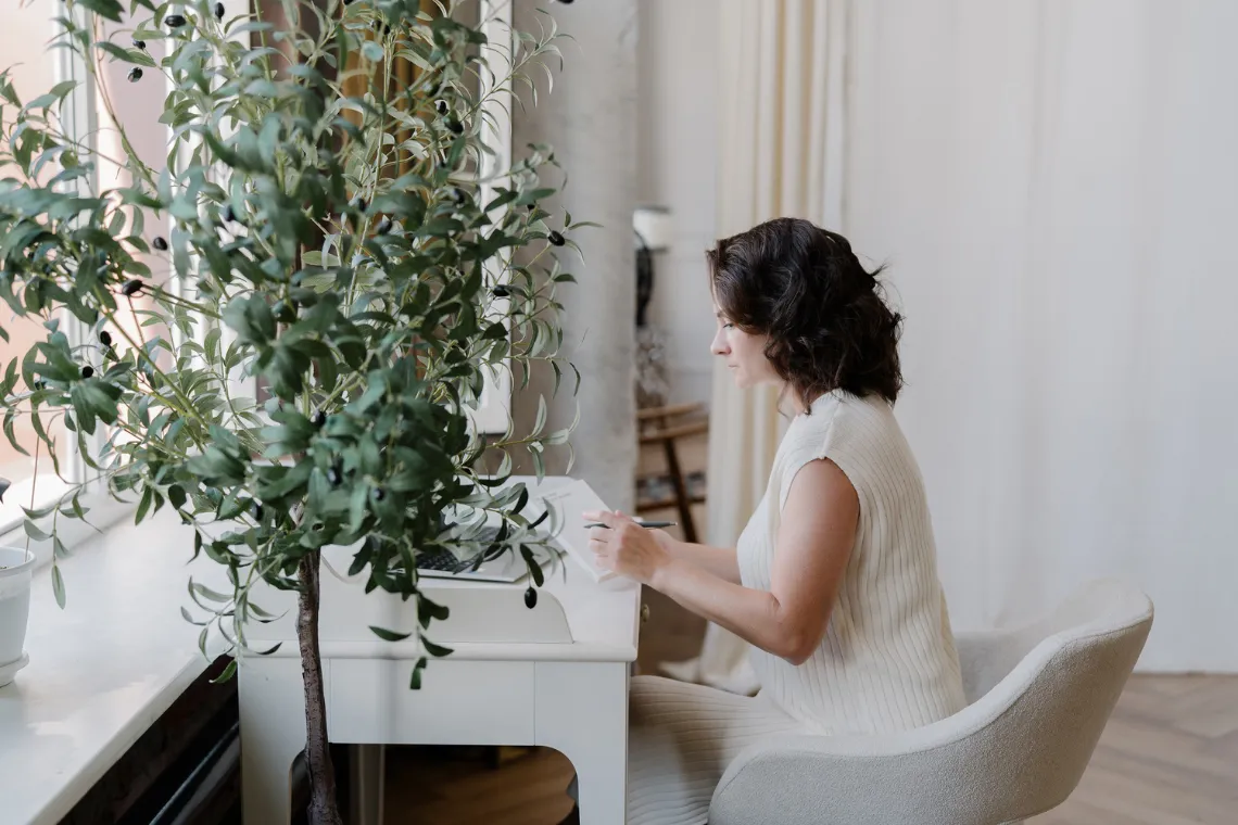 Woman sitting at a desk with a laptop on, writing in a notebook, planning a blogpost.