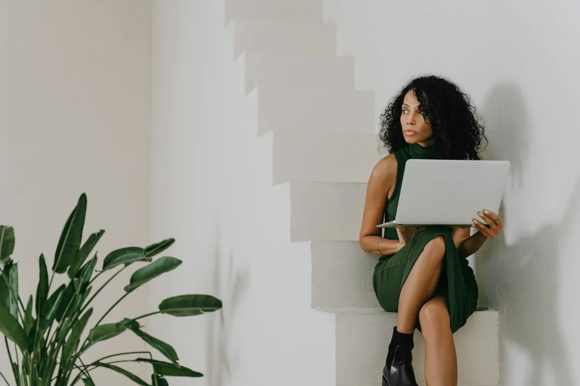 A woman sitting on a staircase with a laptop on her lap, staring into distance.