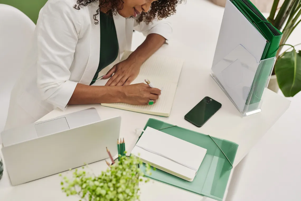Businesswoman sitting at a desk, writing in a notebook with laptop, phone and office supplies nearby.