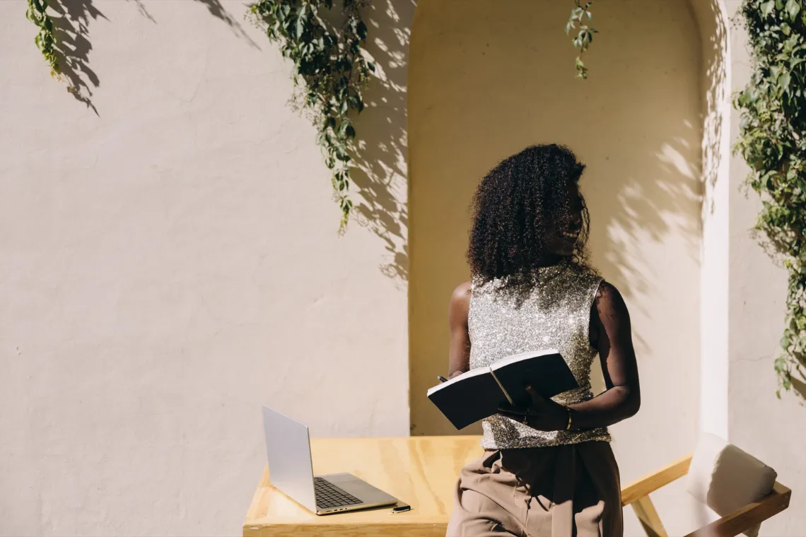 Woman leaning on a table with an open notebook in her hands, laptop open on the table.