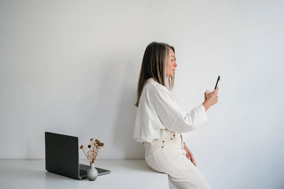 Woman leaning against a desk that has a laptop and flowers on top of it, holding a phone in her hands.