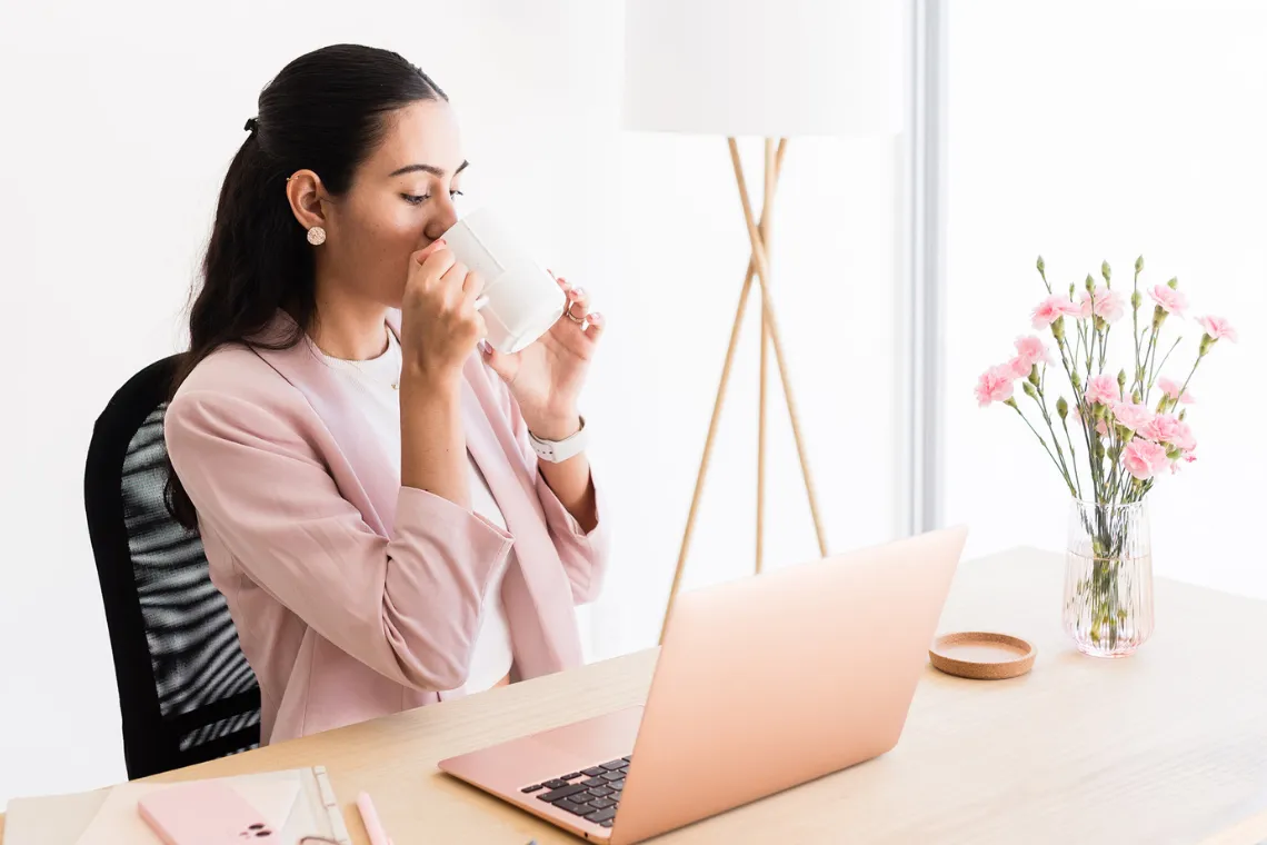 Woman sitting at a table drinking coffee with an open laptop in front of her.