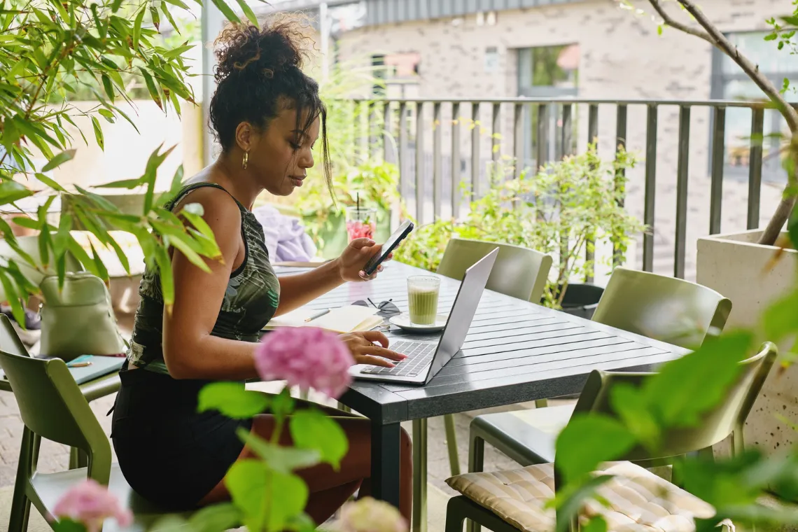 A businesswoman sitting at a garden table surrounded by plants, laptop on the table and a phone in her hand.
